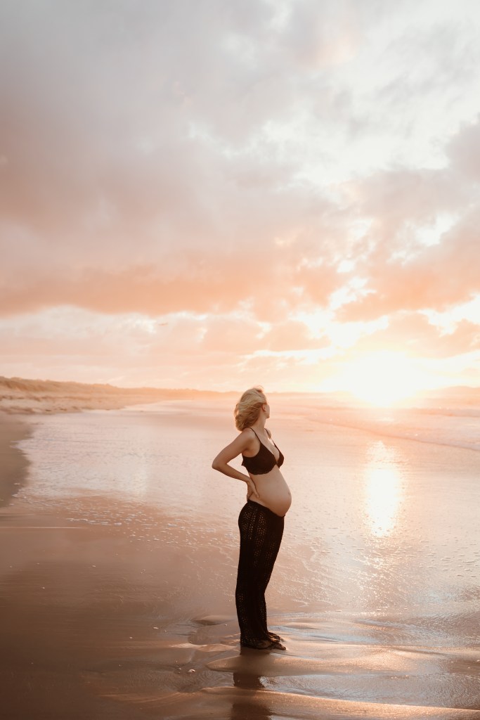 Pregnant blonde woman in black bikini and crochet pants standing on beach with dramatic pink and orange sunset bump session