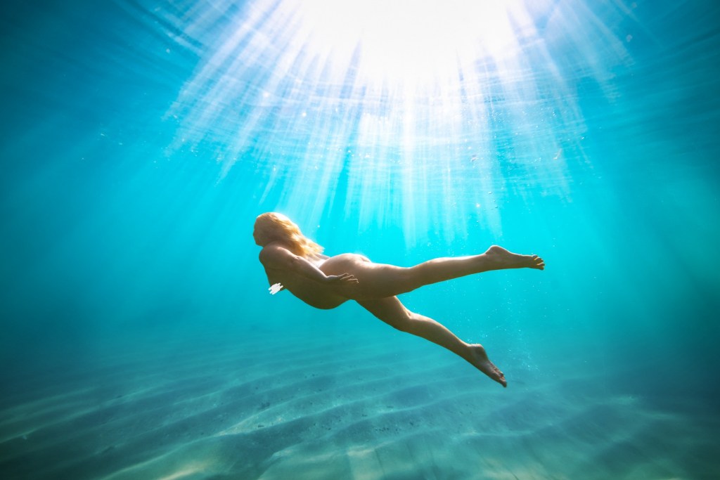Pregnant woman cradling baby bump underwater with brilliant sun rays streaming through turquoise water