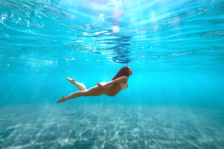 Pregnant woman stretched out underwater with sun flare in deep blue ocean