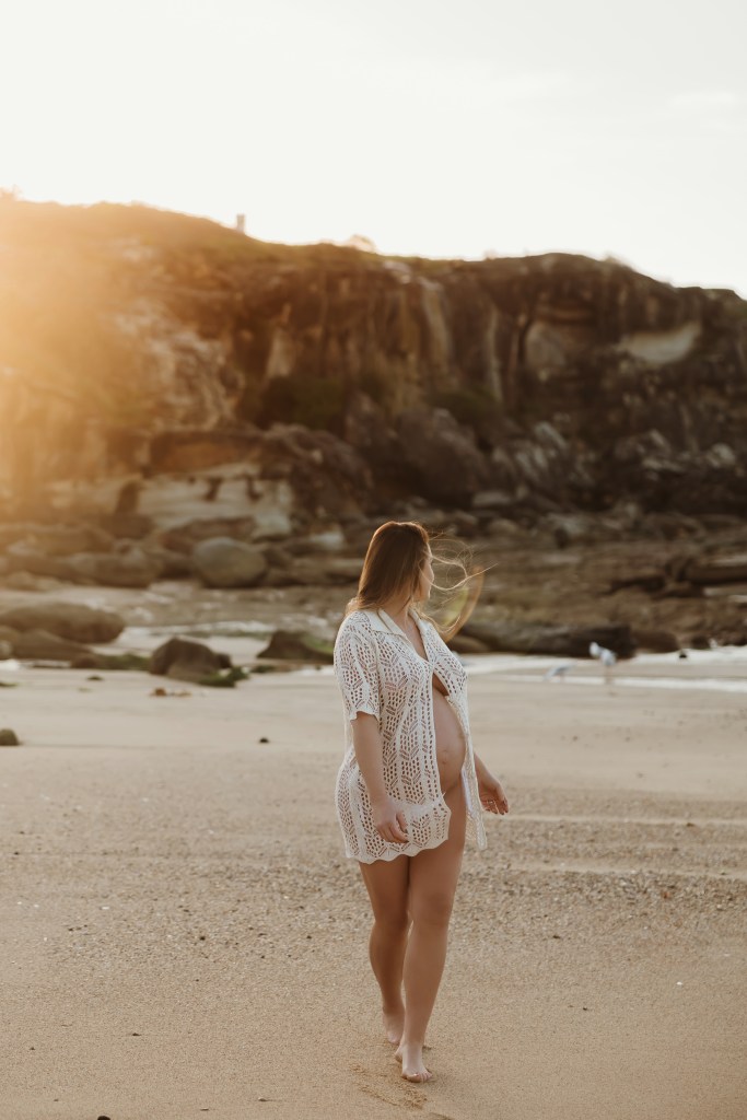 Pregnant woman in white crochet cover-up walking on beach with rocky cliffs at golden hour portraits