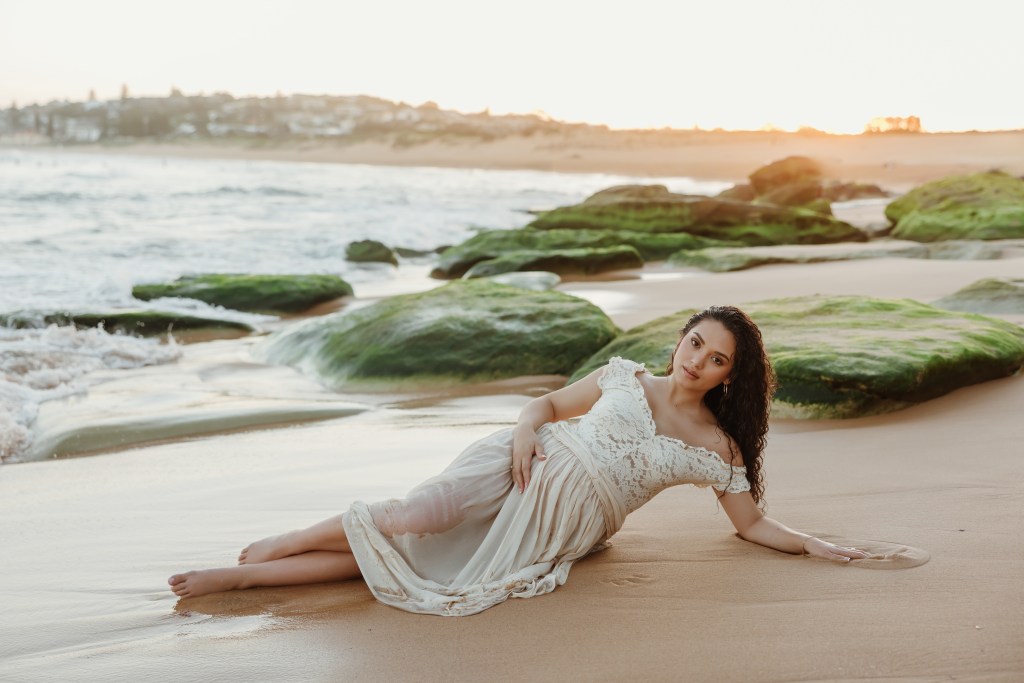 Pregnant woman in white lace flowing dress lying on wet sand with curly hair near mossy rocks photos