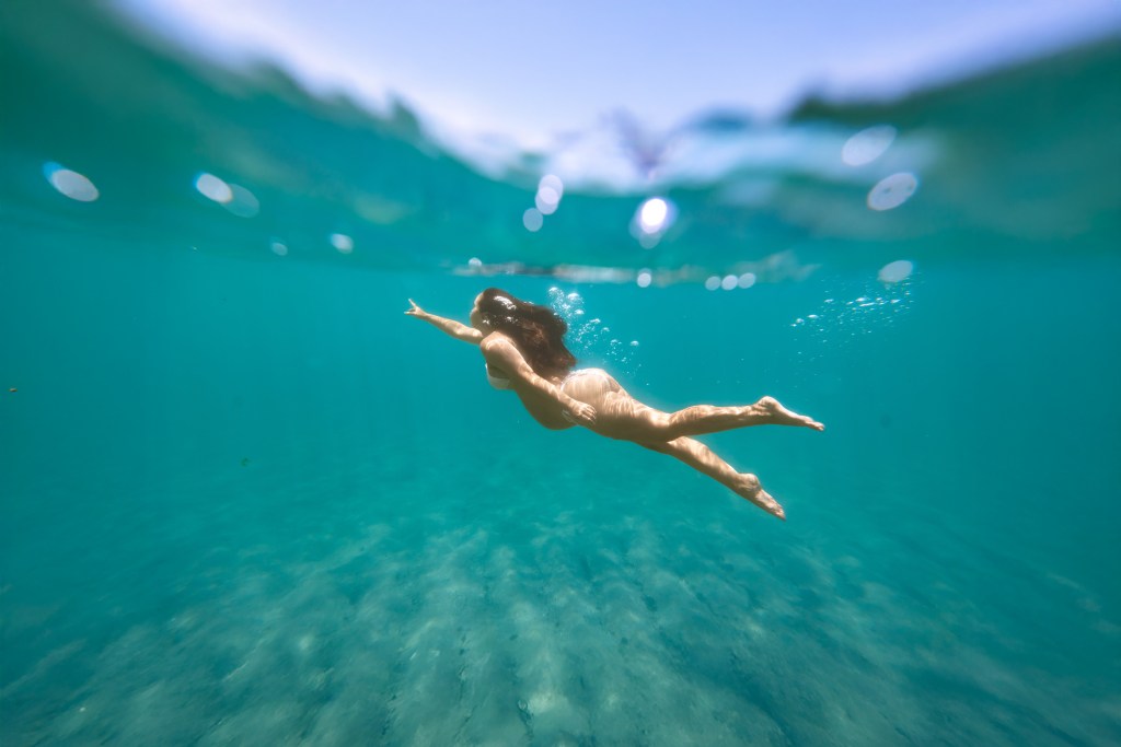 Split-level half-underwater shot of pregnant woman swimming in turquoise ocean with sky visible above the waterline