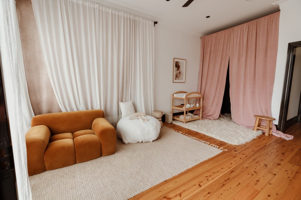 Full room view of newborn photography studio showing mustard couch, white beanbag, open pink velvet drapes, timber floors and rattan decor