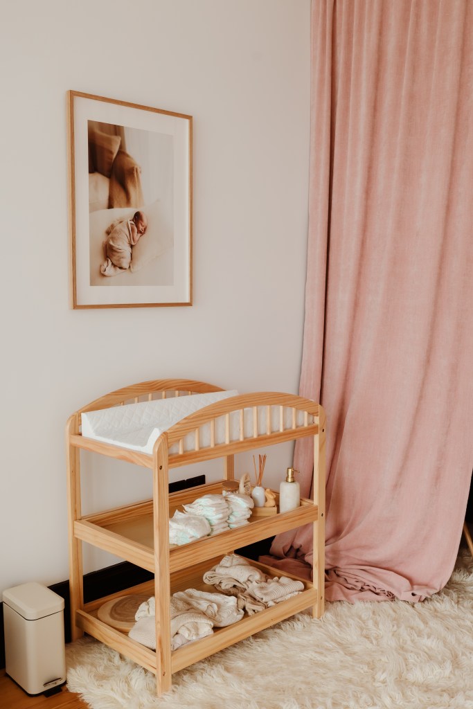 Studio corner detail showing wooden baby change table with nappies and supplies, framed newborn print on wall, pink velvet curtain
